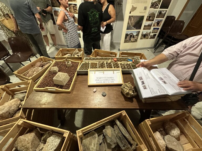 Top view of an interactive display table with dry stone models, a sign reading "Model provided by the Rafel de Cal Nin family," and an open book. Around the table are several wooden boxes filled with different stone samples, and visitors are looking at the information panels in the background.