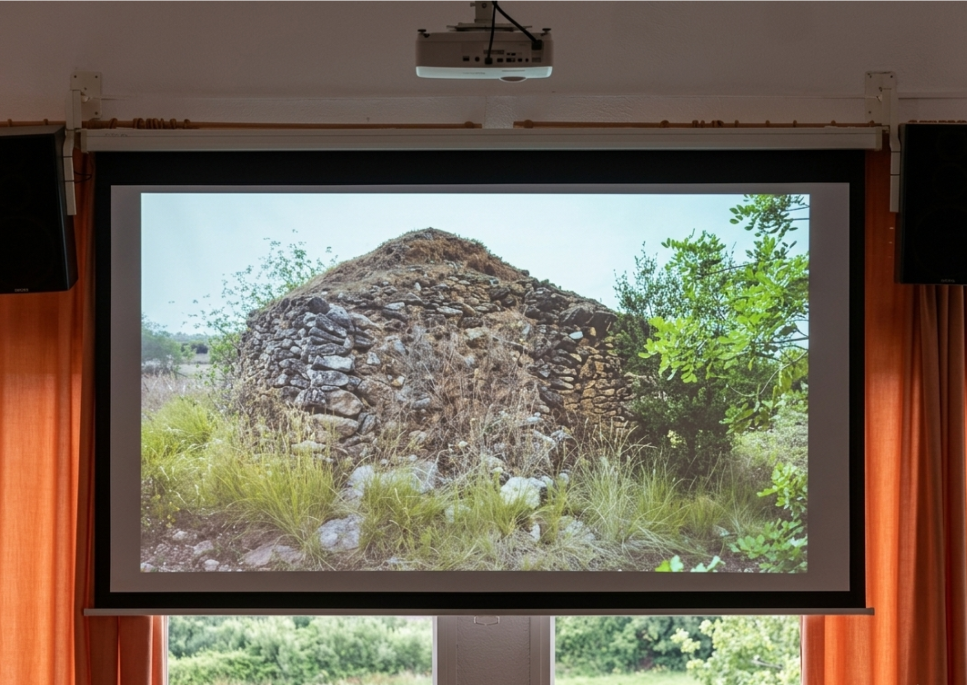 Medium shot of a retractable projection screen flanked by orange curtains and speakers. The screen displays a detailed photograph of a rural cabin built of dry stone amidst wild vegetation.