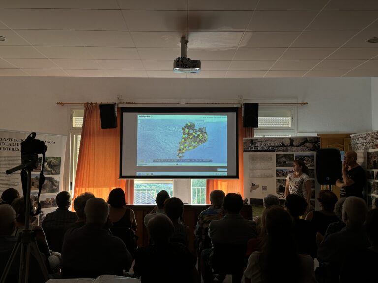 View from the back of a room full of attendees watching a projection on a giant screen. The screen displays an interactive map from Wikipedia, flanked by explanatory banners about the dry stone walling exhibition and two speakers on the right.