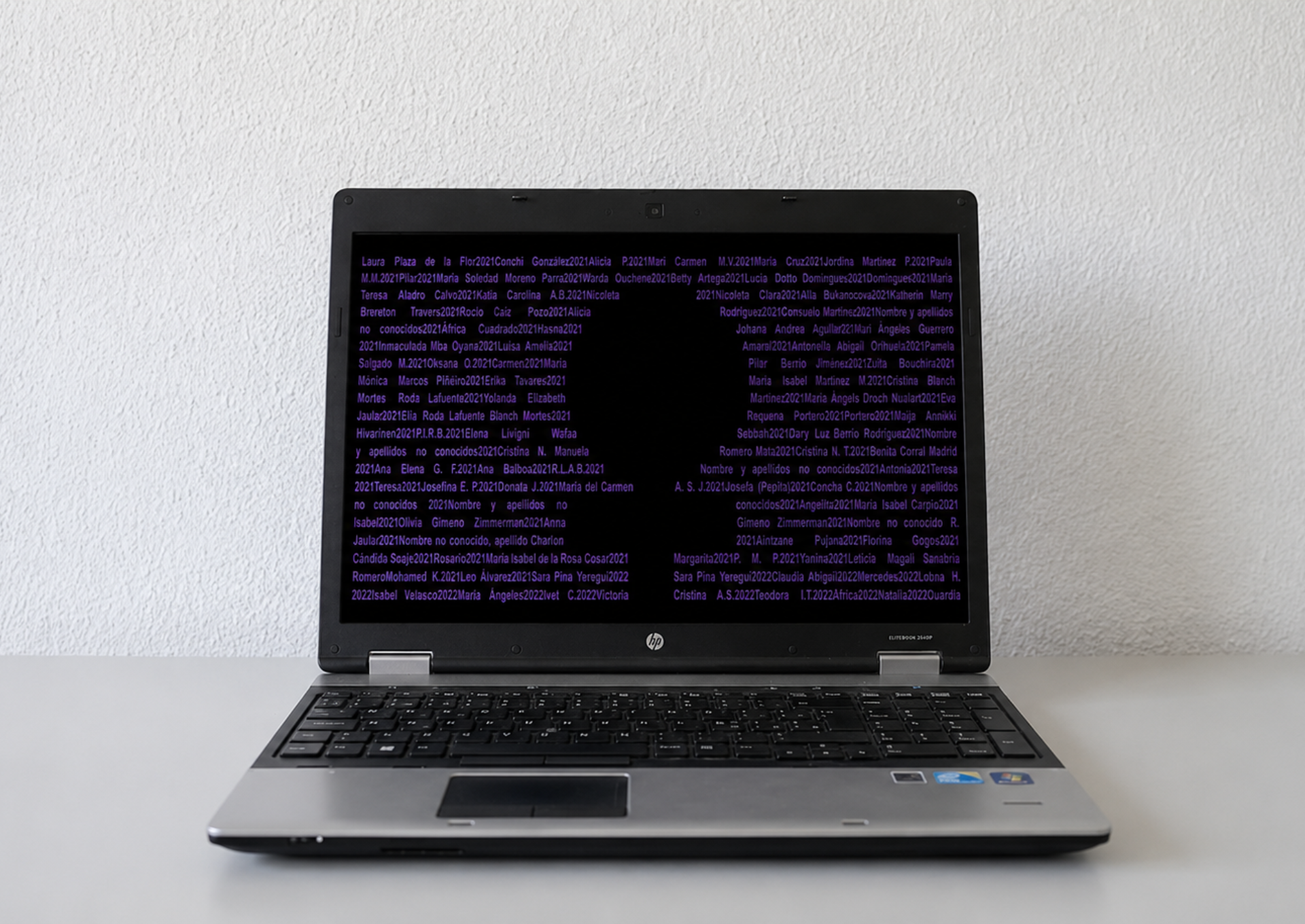 Front view of an open HP laptop against a white background. The black screen displays continuous text in shades of purple, listing the first and last names of women along with their corresponding years, paying tribute to victims of femicide.