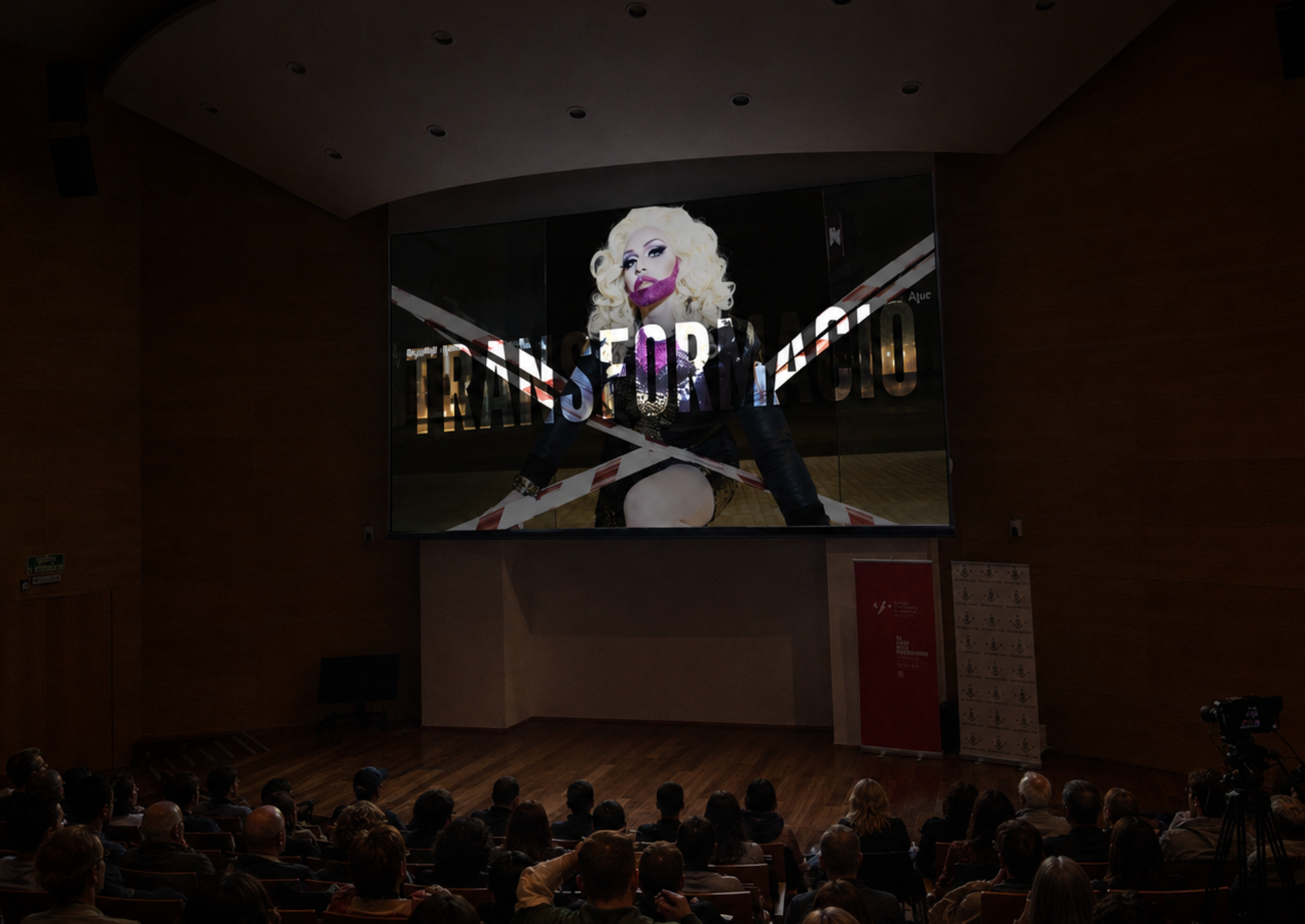 View from the stalls of a darkened auditorium where the public attends the big screen screening of the short film "Transformació", showing the title superimposed on a blonde Drag Queen artist with pink makeup.